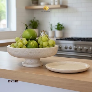 Puede incluir: Un frutero de piedra beige lleno de uvas verdes y limas, con un plato ovalado a juego al lado. El cuenco mide 25 cm de ancho y 10 cm de alto. El entorno de la cocina incluye una encimera de madera y una estufa de acero inoxidable.