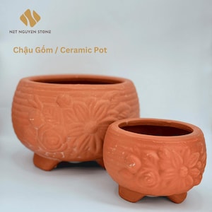 May include: Two terracotta ceramic pots with floral designs. The larger pot is approximately 10 inches in diameter, and the smaller pot is about 6 inches in diameter. Both pots have a rounded shape and sit on three small feet. The text "Chậu Gốm / Ceramic Pot" is visible.