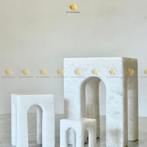 May include: Three white marble accent tables of varying sizes. The tables have a minimalist design with arched legs and a rectangular top. The tables are arranged in a group, showcasing their different heights and shapes.