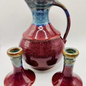 Puede incluir: Un juego de cerámica hecho a mano que incluye una jarra burdeos con una parte superior y un asa esmaltadas en azul y verde, y dos candelabros a juego. Los candelabros tienen una base burdeos y un borde azul y verde.