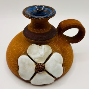 May include: A handmade ceramic pitcher with a handle. The pitcher is primarily brown with a white flower design on the front. The rim is blue, and a dark brown band encircles the neck. The pitcher is approximately 5 inches tall.