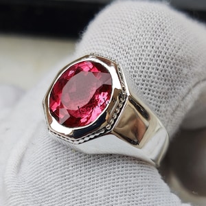 May include: A silver ring with a large, oval-cut, ruby-red gemstone. The ring has a wide band and a detailed bezel setting. The gemstone is the focal point, catching the light and reflecting a vibrant color. The ring is displayed on a white surface.