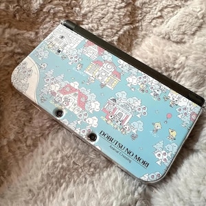 May include: A Nintendo 3DS handheld game console featuring a light blue cover with a cartoon village design. The design includes houses, trees, and characters from "Animal Crossing". The text "DOBUTSU NO MORI Animal Crossing" is printed on the cover.