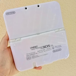 May include: A white Nintendo New 3DS LL handheld gaming console, open to reveal the interior. The console has a glossy finish and features the Nintendo logo. The back of the console displays text including "new: NINTENDO 3DS LL" and "MADE IN CHINA".