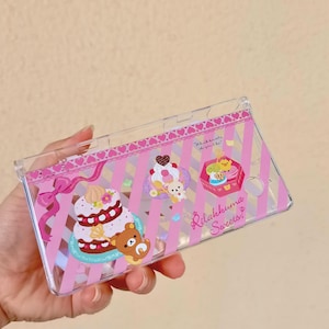 May include: Clear phone case with a pink and white striped background, featuring illustrations of cakes, bears, and the text "Rilakkuma Sweets!" and "Which sweets do you like?" The case has a pink bow and heart accents.