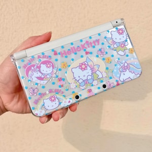May include: A white handheld gaming console with a clear plastic cover featuring Hello Kitty designs. The cover has illustrations of Hello Kitty as a fairy and a unicorn, along with the text "Hello Kitty" in pink. The design includes flowers and butterflies.