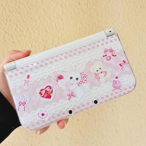 May include: A white and pink Nintendo 3DS case featuring a cute bunny, teddy bear, and heart designs. The case has a polka dot pattern and decorative lace trim. The case is held in a hand.