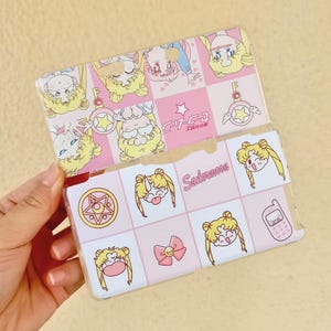 Puede incluir: Una funda de plástico transparente con un diseño rosa y blanco con personajes de Sailor Moon. La funda tiene un patrón de cuadrícula con varias ilustraciones de Sailor Moon, incluyendo la cara del personaje, un lazo y un teléfono.