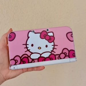 Puede incluir: Una consola de juegos portátil rosa con un gráfico de Hello Kitty en la cubierta frontal. El diseño incluye al personaje blanco de Hello Kitty con un lazo rosa, rodeado de patrones florales rosa oscuro y dos lentes de cámara en el borde superior.