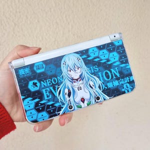 May include: A handheld gaming device featuring a blue and white design with anime-style artwork. The device has a blue and white color scheme with the words "NEON EV" and Japanese text. The artwork depicts a female character with blue hair.