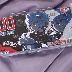 May include: Clear plastic phone case featuring a manga-style illustration of a character with blue hair. The case has the text "00 EVA UNIT" in red and white, along with Japanese text and the name "REI AYANAMI".