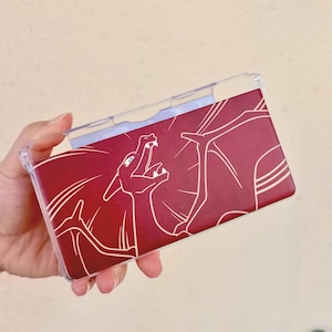 May include: A clear plastic case with a burgundy background featuring a white line drawing of a dragon. The dragon is depicted with its mouth open, and the case is designed to fit a handheld gaming device.