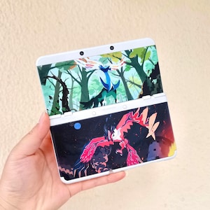 May include: White Nintendo 3DS handheld game console with a custom design featuring a forest scene with a blue and black creature on the top screen and a red and black creature on the bottom screen. The console is open and held in a hand.