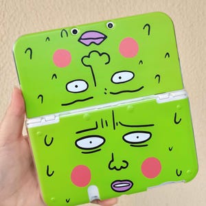 May include: A bright green handheld gaming console case with a cartoon face design. The case is split into two sections, each with a different face. The faces have pink cheeks, black outlines, and various expressions.