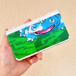 May include: A white Nintendo 3DS handheld game console with a custom skin featuring a blue and green cartoon character. The character has a large mouth and is set against a green background.