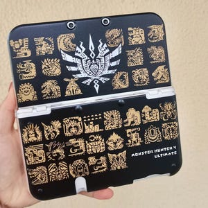 May include: A black handheld gaming console featuring a white emblem and numerous gold-colored creature icons printed on the exterior. The text Monster Hunter 4 Ultimate is printed in white on the lower right corner of the device casing.