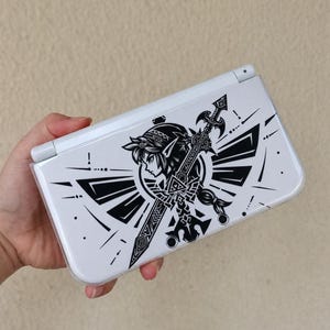 Puede incluir: Una consola de juegos portátil blanca con un estampado gráfico negro en la cubierta superior. El diseño muestra el perfil estilizado de un personaje con orejas puntiagudas, situado detrás de una gran espada y un emblema alado con líneas.