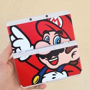 May include: A red and white Nintendo 3DS handheld game console featuring a large graphic of the character Mario. The design includes Mario's face, hat, and overalls. The console is open, revealing the screen and buttons.