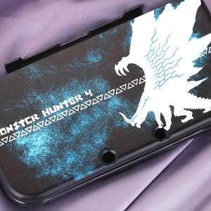 Puede incluir: Funda negra para Nintendo 3DS con un gráfico blanco y azul de un monstruo y el texto "MONSTER HUNTER 4". La funda tiene un acabado mate sobre un fondo morado.