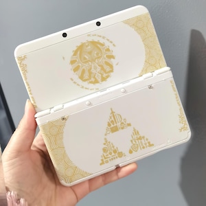 May include: A white Nintendo 3DS handheld gaming console with gold-colored decorative designs. The top cover features a circular emblem, while the bottom cover displays a triangle design. The console is held in a hand.