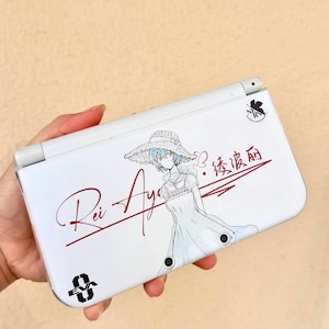 May include: White handheld gaming console with a Rei Ayanami illustration and the text "Rei Aya". The console also features Japanese text and a black logo.