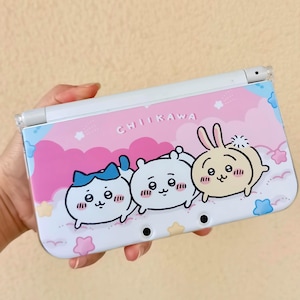May include: A white handheld gaming console featuring a colorful design with the word "CHIIKAWA" and three cartoon characters. The design includes pink clouds, stars, and a blue and pink color scheme.