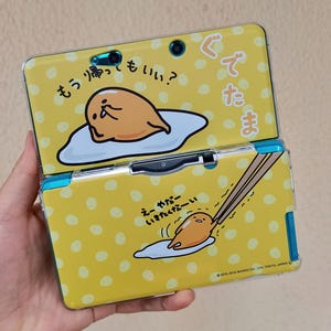 May include: A yellow handheld gaming console featuring a Gudetama character design with white polka dots. The top screen area shows the character with Japanese text, and the bottom area shows the character being picked up by chopsticks with Japanese text.
