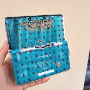 May include: A teal Nintendo 3DS handheld game console with a clear protective case. The case is decorated with a pattern of small black shapes and a group of cartoon characters. The console is open, revealing the screen and buttons.