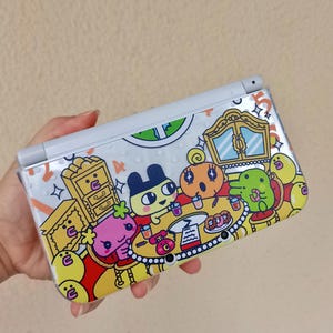 May include: A white handheld gaming console featuring a clear protective case with a colorful graphic print of cartoon characters sitting around a table with furniture, a red telephone, and various food items against a light background.
