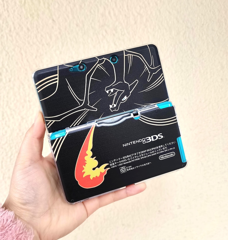 May include: Black Nintendo 3DS handheld game console with a custom skin featuring a white outline of a dragon and a flame graphic. The console has blue accents and the text "NINTENDO 3DS".