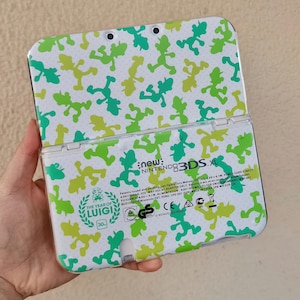 May include: A white New Nintendo 3DS XL handheld game console featuring a repeating green and lime green silhouette pattern of Luigi. The back includes the text New Nintendo 3DS XL and a Year of Luigi 30th anniversary emblem with regulatory text.