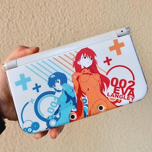 May include: White Nintendo 3DS handheld game console featuring anime characters in blue and orange. The console has a design with plus signs and the text "002 EVA LANGLEY".