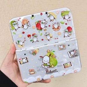 May include: A light gray Nintendo 3DS handheld game console with a decorative design featuring cartoon characters, fruit, and polka dots. The console is open, revealing the screen and buttons. The design includes the character Pochacco.