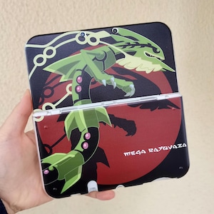 Op de afbeelding: Een draagbare spelcomputer met een zwart en rood grafisch ontwerp van het personage Mega Rayquaza. De console toont de tekst Mega Rayquaza in witte letters tegen een donkere achtergrond met een groot rood cirkelvormig element.