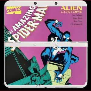 May include: A white handheld gaming console featuring a graphic cover of The Amazing Spider-Man Alien Costume comic book. The cover art displays a black-suited Spider-Man crouching on a building against a pink background with comic text.