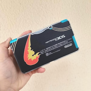May include: Black Nintendo 3DS console with a decorative design featuring a red and yellow flame-like graphic. The console has blue accents and the Nintendo 3DS logo. The text on the console includes Japanese characters and the Nintendo logo.