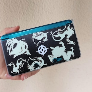 May include: A teal handheld gaming console featuring a black protective skin with a white graphic pattern of multiple dragons and a central circular emblem. The device has a clear outer shell and visible camera lenses on the back surface.
