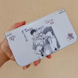 Puede incluir: Una consola de juegos portátil blanca con una ilustración de líneas negras de cuatro personajes de anime. El texto dice "THIRD", "FIRST", "SECOND", "FIFTH", "DANGER" y "NERV" con un logotipo de hoja. El dispositivo tiene dos lentes de cámara circulares.