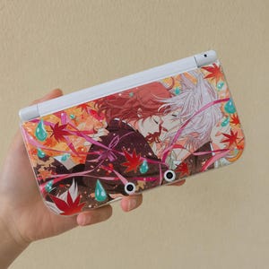 May include: A white Nintendo 3DS handheld game console with a custom cover featuring an anime-style illustration. The cover displays two figures embracing, surrounded by autumn leaves and water droplets in shades of red, orange, and teal.