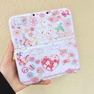 May include: A white handheld gaming console case with a decorative floral and animal design. The case features a rabbit, butterflies, flowers, and a heart-shaped design in pink and red. The case is open, revealing the screen and buttons.