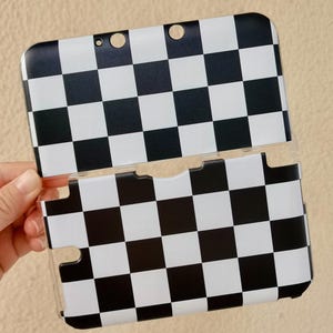 May include: A protective hard shell case for a handheld gaming console featuring a high-contrast black and white checkered pattern. The case consists of two separate pieces with precise cutouts for buttons, ports, and camera lenses.