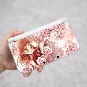 Pode incluir: Consola de jogos portátil branca com uma capa transparente com uma ilustração de anime. A imagem mostra um casal sob flores de cerejeira rosa, com um homem beijando uma mulher.