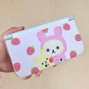 May include: A light pink Nintendo 3DS case featuring a cartoon bear in a pink bunny costume and a yellow bear, both holding strawberries. The case is decorated with scattered strawberries.