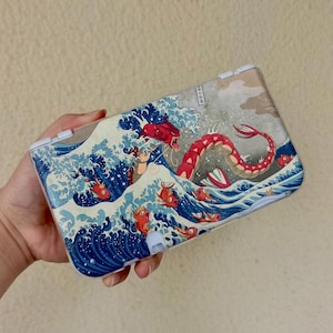 May include: A handheld gaming console featuring a graphic print inspired by The Great Wave off Kanagawa, depicting a large red serpentine creature and several smaller orange fish swimming amidst stylized blue and white ocean waves.