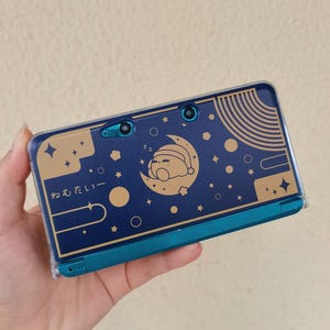 Può includere: Una console di gioco portatile blu scuro con una grafica dorata che raffigura un personaggio che dorme su una luna crescente, circondato da stelle e pianeti. Il design include il testo giapponese "ねむたい" e una custodia protettiva trasparente.