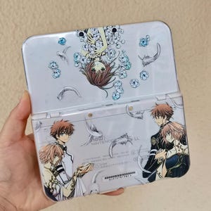May include: A white New Nintendo 3DS LL handheld gaming console featuring a clear protective cover decorated with anime-style illustrations of characters and floating feathers. The top screen area shows a character surrounded by blue flowers.