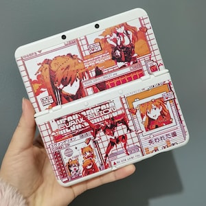 Op de afbeelding: Witte Nintendo 3DS handheld gameconsole met een aangepast ontwerp met illustraties in anime-stijl in rood, oranje en wit. Het ontwerp bevat tekst en pixelachtige graphics, met de woorden "EVANGELION" en Japanse karakters zichtbaar.