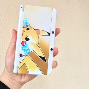 May include: White handheld gaming console featuring a Pikachu design. The yellow and brown Pikachu illustration is on the front of the console, with blue lightning bolts. The console has black buttons and a white stylus.