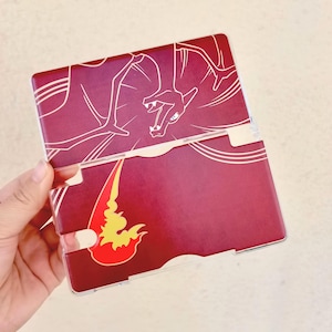 May include: A burgundy and clear plastic case featuring a white outline of a dragon on the top half. The bottom half has a flame design in red and yellow. The case is designed to hold a gaming device.