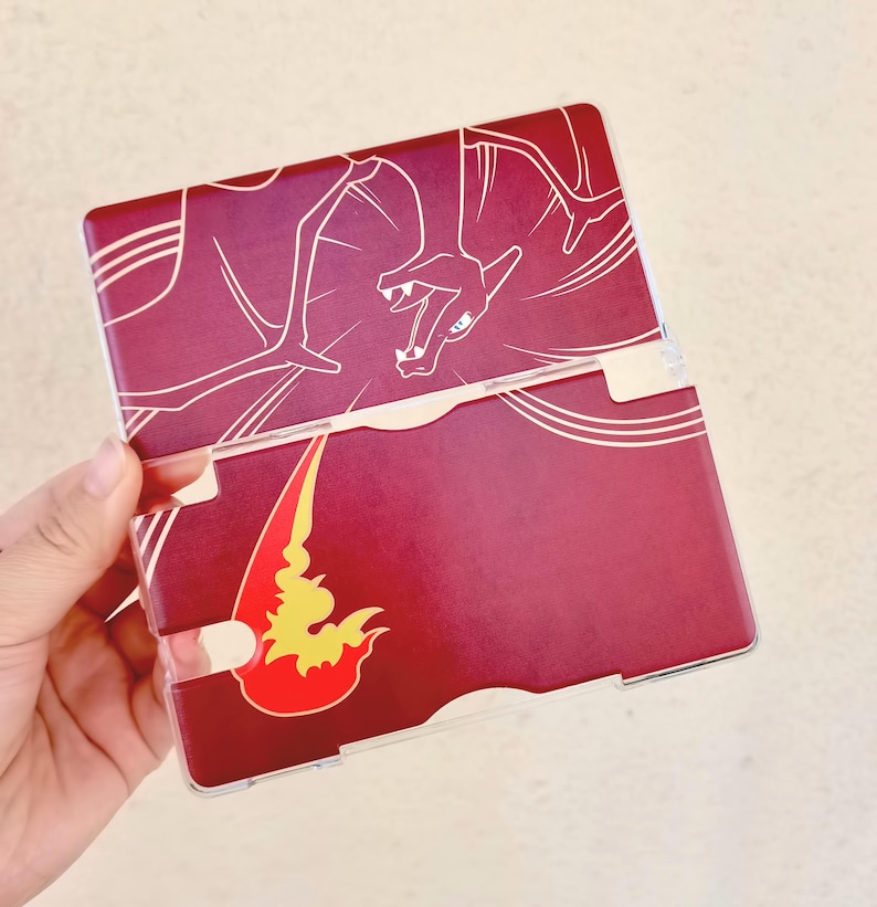 May include: A burgundy and clear plastic case featuring a white outline of a dragon on the top half. The bottom half has a flame design in red and yellow. The case is designed to hold a gaming device.